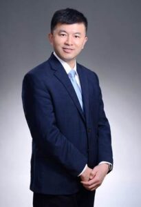 Image of a smiling Chinese man dressed in a navy blue business suit and light blue tie.